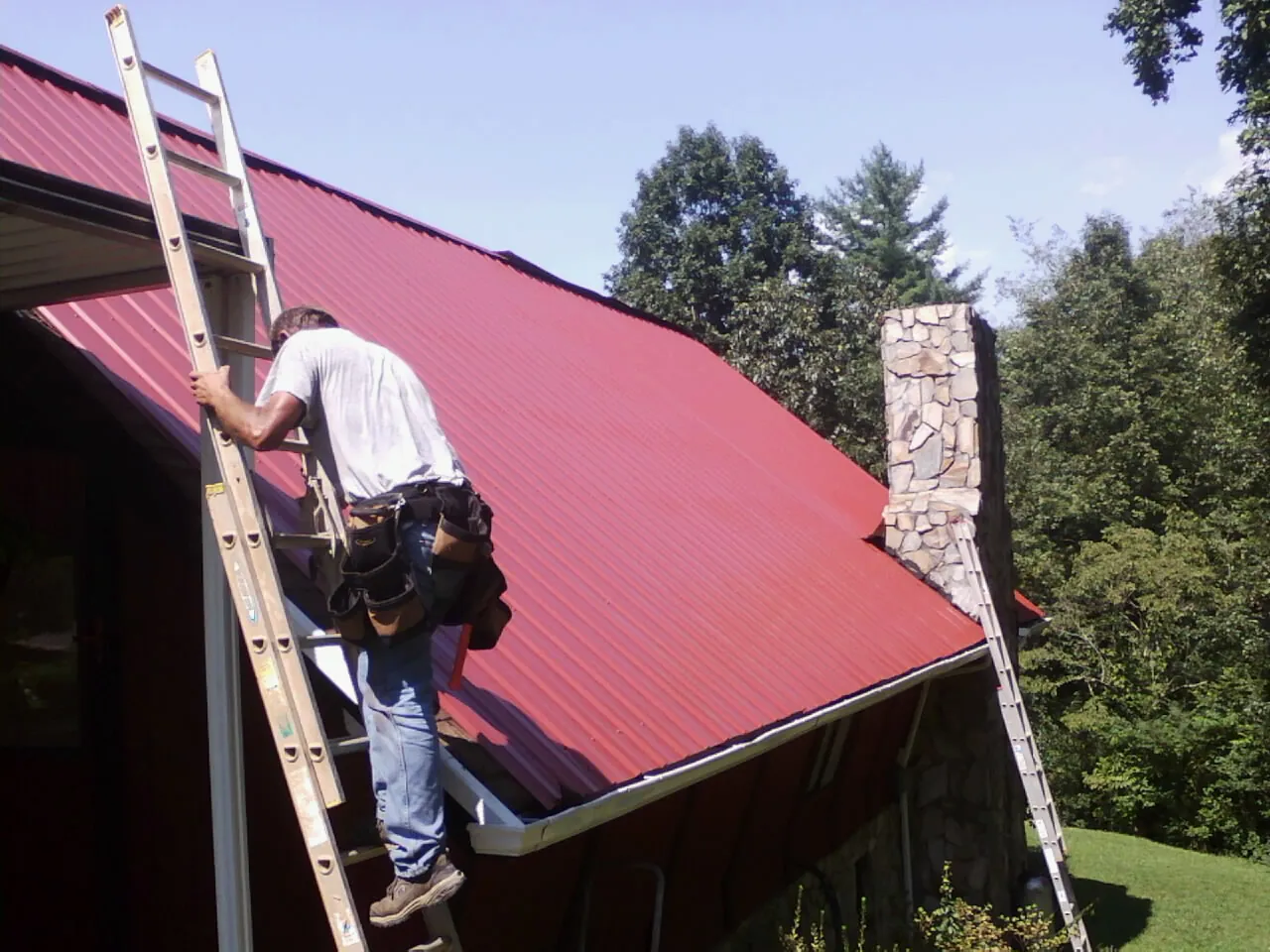 Roofer on a ladder beside a red metal roof and stone chimney — Soffit & Fascia Repair by Roofing Brothers in Harrellsville